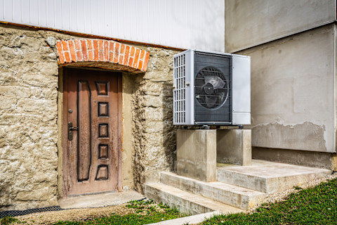 Dieses Bild zeigt eine moderne Wärmepumpe neben einem alten Steingebäude mit einer verzierten Holztür mit Schnitzereien.