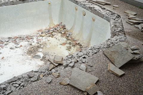 Dieses Bild zeigt ein leeres Schwimmbad, das abgerissen wird. Sowohl am Beckenrand als auch im Inneren liegen Trümmer. Alles ist mit einer Schicht feinen, weißen Staubs bedeckt.