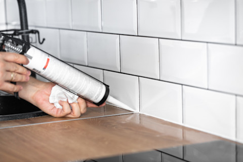 Dieses Bild zeigt ein Paar Hände, die Silikon in einer Tube verwenden, um den Spalt zwischen einer Küchenarbeitsplatte und einer gefliesten Wand abzudichten. In der Hand der Person liegt ein Stück zerknülltes Seidenpapier, um eventuelle Rückstände vom Silikon zu entfernen.