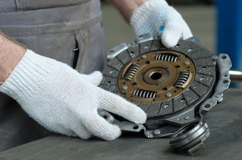 Ein Mechaniker in grauer Arbeitskleidung hält ein Kupplungsteil in den Händen. Die Person trägt weiße Arbeitshandschuhe mit blauen Punkten. Das Bild zeigt den Kupplungsmechanismus, der mit einer Druckplatte und Federn ausgestattet ist. Die Szene deutet darauf hin, dass der Mechaniker dabei ist, die Kupplung eines Fahrzeugs zu reparieren oder auszutauschen. Im Vordergrund liegt ein weiteres Teil, möglicherweise ein Ausrücklager, das ebenfalls mit der Kupplung zusammenhängt.