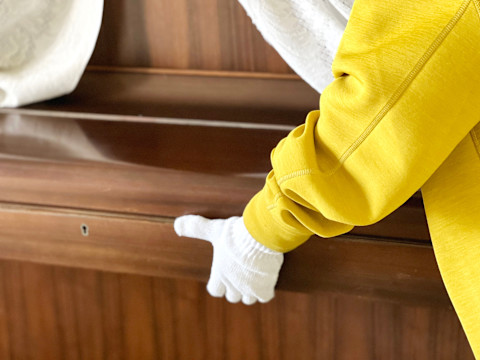 Dieses Bild zeigt eine Person in einem gelben Overall und weißen Stoffhandschuhen, die vorsichtig ein Klavier bewegt. Die Klappe ist heruntergeklappt und schützt die Tasten.