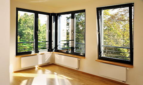 Ein heller Raum mit großen, schwarzen Fensterrahmen und einer herrlichen Aussicht auf einen grünen Garten. Die Fenster lassen viel Tageslicht herein, das den Raum warm und einladend macht. Unter den Fenstern befinden sich weiße Heizkörper und Fensterbänke aus Holz. Der Boden ist mit hellem Holzparkett ausgelegt. Die Wände sind in einem neutralen Farbton gestrichen, der die Helligkeit des Raumes unterstreicht.