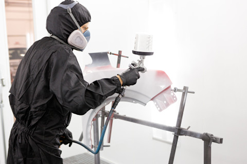 Das Bild zeigt eine Person in Schutzkleidung, die mit einer Spritzpistole Autoteile lackiert. Die Person trägt eine Atemschutzmaske und einen schwarzen Overall, um sich vor den Dämpfen und Chemikalien zu schützen, die beim Lackieren entstehen. Das Autoteil, wahrscheinlich eine Stoßstange, wird in einer speziellen Lackierkabine sorgfältig mit einer gleichmäßigen Farbschicht besprüht. Der Lackierungsprozess erfordert Präzision, damit das Endergebnis makellos und professionell ist.