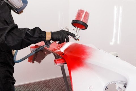 Das Bild zeigt eine Person, die eine Spritzpistole verwendet, um eine rote Lackierung auf eine Autostoßstange aufzutragen. Der Arbeiter trägt eine Schutzmaske und Handschuhe, um sich vor den Chemikalien des Lackes zu schützen. Der Lackierprozess erfolgt in einer professionellen Umgebung, möglicherweise in einer Werkstatt oder einer Lackierkabine. Der rote Farbton hebt sich deutlich von der weißen Grundierung ab und wird gleichmäßig auf die Oberfläche der Stoßstange aufgetragen.