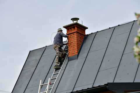 Dieses Bild zeigt einen Mann, der eine Leiter auf das Dach des Hauses klettert, um den Schornstein zu reparieren. Der Schornstein besteht aus rot/orangefarbenen Ziegeln und das Dach ist dunkelgrau.