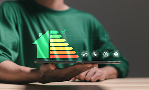 Das Bild zeigt eine Person, die ein Tablet hält, auf dem ein Energielabel in Form eines Hauses angezeigt wird. Die Energieeffizienzskala reicht von "A" (grün und effizient) bis "G" (rot und ineffizient). Auf der Skala wird der Energieverbrauch eines Hauses oder Geräts visuell dargestellt. Zusätzlich sind Symbole zu sehen, die nachhaltige Aspekte wie Umweltschutz, erneuerbare Energien, Kosteneinsparungen und CO2-Reduktion symbolisieren. Dies betont die Bedeutung von Energieeffizienz im modernen Leben.