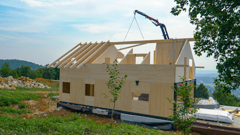 Das Bild zeigt ein Holzfertighaus, dass mit Hilfe eines Krans in einer Naturlandschaft aufgestellt wird.