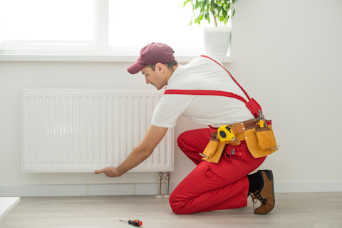 Dieses Bild zeigt einen Heizungsbauer, der einen Heizkörper installiert, um ein Haus mit Wärme zu versorgen. Der Ingenieur trägt einen roten Overall und einen Werkzeuggürtel mit den für die Arbeit notwendigen Werkzeugen.