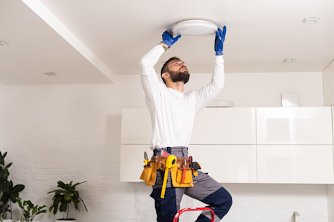 Dieses Bild zeigt einen Handwerker, der eine Deckenleuchte installiert. Er steht auf einer Leiter, um die Armatur erreichen zu können.
