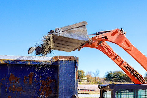 Ein Bagger ist dabei, ein Gebäude abzureißen. Der Greifarm des Baggers hebt Schutt und Trümmer, während dichte Staubwolken die Szene umgeben. Zerbrochene Ziegelsteine und Betonstücke liegen verstreut auf dem Boden. Im Hintergrund sind Teile der beschädigten Gebäudestruktur sichtbar. Die Aufnahme vermittelt die Dynamik und die Kraft des Abrissprozesses.