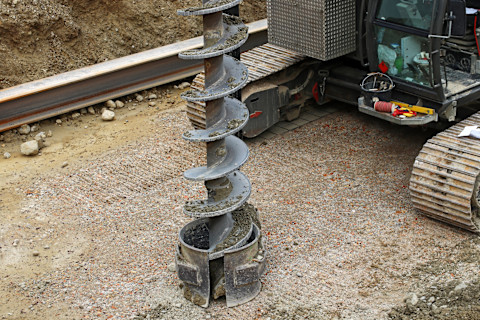 Das Bild zeigt eine große Bohrmaschine, die auf einer Baustelle verwendet wird. Es handelt sich um einen schweren Erdbohrer, der zum Bohren von Löchern im Boden dient, wahrscheinlich für Fundamente oder Pfähle. Der Erdbohrer ist mit einer Schraubenform versehen, die es ermöglicht, Erde aus der Tiefe an die Oberfläche zu transportieren. Im Hintergrund ist das Kettenfahrzeug zu sehen, das den Bohrer antreibt, sowie einige Werkzeuge und Ausrüstungsgegenstände, die auf der Baustelle verwendet werden.