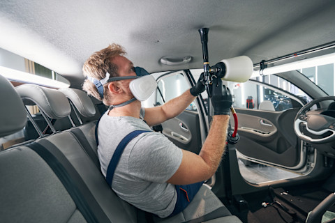 Das Bild zeigt einen Mann mit Overall und Schutzmaske, der den Dachhimmel eines Autos professionell reinigt.