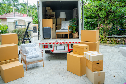 Ein Umzugswagen steht offen auf einer Auffahrt, umgeben von mehreren Umzugskartons und Möbeln, die teilweise verpackt sind. In dem LKW befinden sich verschiedene Gegenstände, darunter ein Sessel, eine Pflanze und ein Koffer. Kartons und Möbelstücke stehen ordentlich gestapelt bereit, um in das Fahrzeug geladen zu werden. Die Szene deutet auf einen Umzug hin, der entweder begonnen hat oder kurz vor dem Abschluss steht. Im Hintergrund ist ein gepflegter Garten mit Bäumen und Büschen zu sehen, was auf eine ruhige Wohngegend hinweist.
