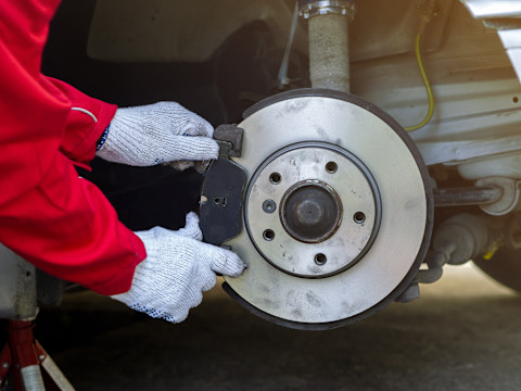 Eine Nahaufnahme zeigt die Hände einer Person, die weiße Arbeitshandschuhe und einen roten Arbeitsoverall trägt, während sie an einer Bremsscheibe arbeitet. Die Bremsscheibe ist an einem Fahrzeug montiert und hat eine silberne, metallische Oberfläche mit einigen leichten Gebrauchsspuren. Die Person hält eine Bremsbelagplatte, die an der Seite der Bremsscheibe befestigt wird. Im Hintergrund sind Teile der Fahrzeugaufhängung und ein gelbes Kabel zu sehen.
