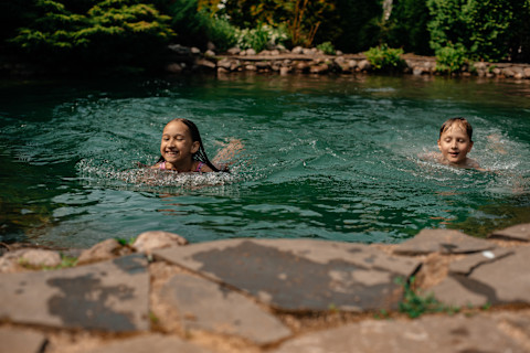 Das Bild zeigt zwei Kinder, die in einem Schwimmteich schwimmen.