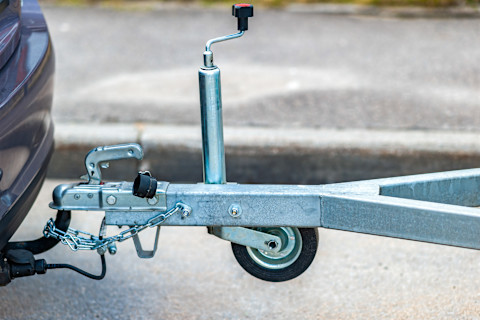 Nahaufnahme einer Anhängerkupplung, die an einem Fahrzeug befestigt ist. Das Bild zeigt den vorderen Teil eines Anhängers mit einer silbernen Metallkupplung, die am Heck eines Fahrzeugs angebracht ist. Die Kupplung ist durch eine Sicherheitskette gesichert. An der Anhängerkupplung ist ein Stützrad mit einer Kurbel zur Höhenverstellung montiert. Der Untergrund ist asphaltiert, und im Hintergrund ist ein Bordstein zu sehen.