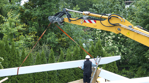 Das Bild zeigt einen Mann in Arbeitskleidung und Schutzhelm, der mithilfe eines Krans einen Balkon konstruiert. Im Hintergrund befindet sich ein Wald.