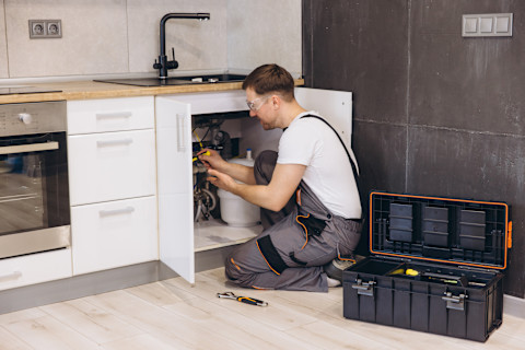Das Bild zeigt einen Mann in Arbeitskleidung und Schutzbrille, der eine Küchenspüle installiert. Im Vordergrund sieht man eine Box mit Arbeitsmaterialien.