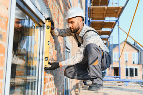 Das Bild zeigt einen Bauarbeiter mit Helm und Overall, der mit einer Wasserwaage testet, ob ein neues Fenster richtig eingebaut ist.