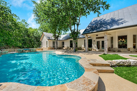 Das Bild zeigt ein wunderschönes Haus mit einem großen, klaren Swimmingpool im Vordergrund. Das Haus hat eine moderne Architektur mit einem hellen Außenanstrich und einem metallischen Dach. Rund um den Pool gibt es einen gepflegten Garten mit Bäumen, die angenehmen Schatten spenden. Die Terrasse lädt zum Entspannen ein, und es ist ein perfekter Ort, um warme Sommertage zu genießen. Die blau schimmernde Wasseroberfläche reflektiert den sonnigen Himmel, was dem Bild eine einladende und entspannte Atmosphäre verleiht.