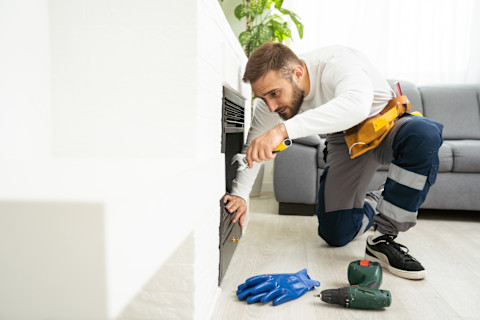 Das Bild zeigt einen Mann in Arbeitskleidung, der einen Kaminofen installiert. Im Vordergrund sieht man eine Bohrmaschine und ein paar Schutzhandschuhe.