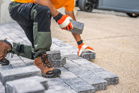Das Bild zeigt einen Bauarbeiter, der Pflastersteine verlegt. Der Arbeiter trägt robuste Arbeitsstiefel, Handschuhe und eine Arbeitshose, um sich vor Verletzungen zu schützen. Die Pflastersteine werden präzise verlegt, um eine stabile und gleichmäßige Fläche zu schaffen. Dies könnte eine Terrasse, eine Einfahrt oder ein Gehweg sein. Die sorgfältige Arbeit trägt zu einem langlebigen und ästhetisch ansprechenden Ergebnis bei.