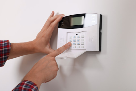 Das Bild zeigt eine Person, die eine Alarmanlage bedient. Die Hände des Nutzers geben einen Code auf dem Tastenfeld ein, um das Sicherheitssystem entweder zu aktivieren oder zu deaktivieren. Solche Systeme werden häufig zum Schutz von Häusern oder Büros vor Einbruch verwendet. Die Person trägt ein Hemd mit einem karierten Muster, und die Alarmanlage ist an einer weißen Wand montiert.