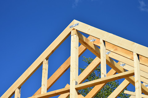 Nahaufnahme eines Dachstuhls im Bau, der aus hellen, unbehandelten Holzbalken besteht. Die Balken sind mit Metallwinkeln und Verbindungselementen verstärkt. Der Dachstuhl hat eine dreieckige Form und ist deutlich vor einem klaren, blauen Himmel sichtbar. Im Hintergrund ist teilweise ein grüner Baum zu erkennen, der auf eine ländliche oder naturnahe Umgebung hinweist. Die Holzkonstruktion steht im Kontrast zum leuchtend blauen Himmel, was auf sonniges Wetter hindeutet.