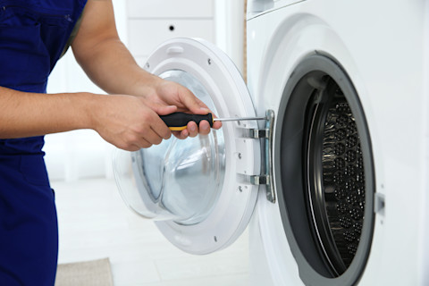 Nahaufnahme eines Technikers, der in blauer Arbeitskleidung eine Waschmaschine repariert. Die Person hält einen Schraubenzieher in der Hand und arbeitet an den Scharnieren der Tür der Frontlader-Waschmaschine. Die Tür ist geöffnet, und die Trommel der Waschmaschine ist sichtbar. Der Schraubenzieher wird verwendet, um die Schrauben an der Tür zu justieren oder zu entfernen. Die Umgebung ist hell, und im Hintergrund sind unscharf weiße Möbel zu erkennen.