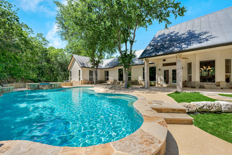 Das Bild zeigt ein wunderschönes Haus mit einem großen, klaren Swimmingpool im Vordergrund. Das Haus hat eine moderne Architektur mit einem hellen Außenanstrich und einem metallischen Dach. Rund um den Pool gibt es einen gepflegten Garten mit Bäumen, die angenehmen Schatten spenden. Die Terrasse lädt zum Entspannen ein, und es ist ein perfekter Ort, um warme Sommertage zu genießen. Die blau schimmernde Wasseroberfläche reflektiert den sonnigen Himmel, was dem Bild eine einladende und entspannte Atmosphäre verleiht.