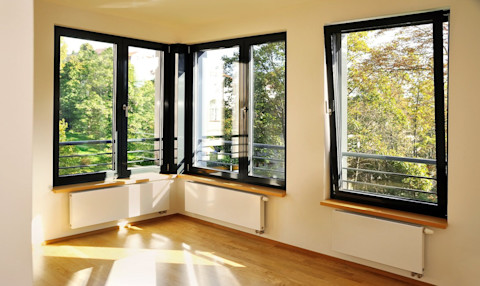 Ein heller Raum mit großen, schwarzen Fensterrahmen und einer herrlichen Aussicht auf einen grünen Garten. Die Fenster lassen viel Tageslicht herein, das den Raum warm und einladend macht. Unter den Fenstern befinden sich weiße Heizkörper und Fensterbänke aus Holz. Der Boden ist mit hellem Holzparkett ausgelegt. Die Wände sind in einem neutralen Farbton gestrichen, der die Helligkeit des Raumes unterstreicht.
