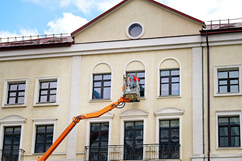 Ein Arbeiter in einer orangefarbenen Arbeitsplattform ist dabei, die Fassade eines mehrstöckigen Gebäudes zu reinigen. Das Gebäude hat eine helle, beigen Fassade mit großen, schwarzen Fensterrahmen. Die Arbeitsplattform ist an einem langen, orangefarbenen Auslegerarm befestigt, der von einem Fahrzeug auf dem Boden gesteuert wird. Der Arbeiter ist in einem Sicherheitskäfig und trägt eine rote Jacke. Der Himmel im Hintergrund ist teilweise bewölkt. Die Szene zeigt typische Fassadenreinigungsarbeiten an einem historischen oder repräsentativen Gebäude.