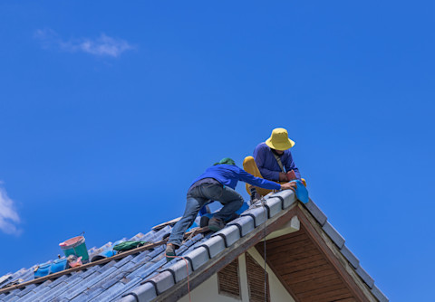 Zwei Arbeiter in blauer Arbeitskleidung und Hüten arbeiten auf dem Dach eines Hauses. Sie sind dabei, Dachziegel an der Spitze des Daches zu installieren oder zu reparieren. Das Dach besteht aus grauen Ziegeln, und die Arbeiter sind mit Sicherheitsseilen gesichert. Der Himmel im Hintergrund ist strahlend blau ohne Wolken, was auf einen sonnigen Tag hindeutet. Die Szene zeigt eine typische Dachdeckerarbeit auf einem Wohnhaus.