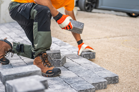 Das Bild zeigt einen Bauarbeiter, der Pflastersteine verlegt. Der Arbeiter trägt robuste Arbeitsstiefel, Handschuhe und eine Arbeitshose, um sich vor Verletzungen zu schützen. Die Pflastersteine werden präzise verlegt, um eine stabile und gleichmäßige Fläche zu schaffen. Dies könnte eine Terrasse, eine Einfahrt oder ein Gehweg sein. Die sorgfältige Arbeit trägt zu einem langlebigen und ästhetisch ansprechenden Ergebnis bei.